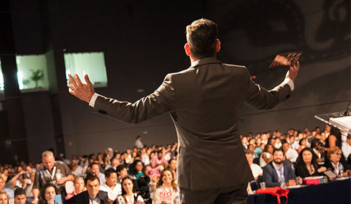 Man on stage, speaking to a large audience with arms outstretched, representing the leadership and communication skills gained from the Advanced Communications Master Practitioner Program